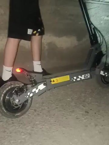 The video shows a person riding an electric scooter in a dimly lit, possibly concrete, environment. The rider is wearing black shorts and white socks with black shoes. The scooter has a red taillight that is illuminated. Dust or dirt is being kicked up from the rear wheel as the scooter moves, suggesting it is on a rough or unpaved surface. The lighting suggests the time is either dusk, night, or the location is indoors without sufficient illumination. The perspective is from behind and to the side of the rider.
