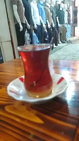 Here is a description of the video:

The video shows a glass of dark red tea with a small spoon in it, sitting on a white saucer with red accents. The saucer rests on a wooden table. In the background, there's a display of mannequins dressed in casual clothing, standing in front of a shop window. The scene appears to be outdoors, possibly at a cafe or shop with outdoor seating. The focus is on the tea, with the mannequins and background providing context to the setting. The lighting is moderately bright, highlighting the color of the tea and the texture of the wooden table.
