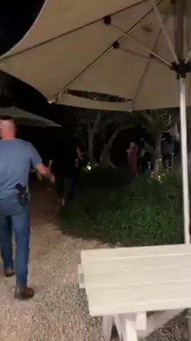 The video shows a person walking towards a gathering of people in an outdoor setting at night. The person is wearing a blue t-shirt, jeans, and brown shoes, and walks on a gravel path under a large beige umbrella. Several other people are visible in the background, some standing and others walking amidst trees and white tables and chairs. The scene is illuminated by artificial lighting, creating a partially lit environment. In the beginning the sound of an air horn can be heard.