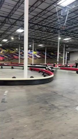 The video shows a go-kart track indoors. There are at least two go-karts racing on the track, both driven by people wearing helmets. The track has red and black barriers along its edges. The building has a high ceiling with exposed beams and lights. The walls are decorated with racing-themed murals and signage. A scoreboard is visible. The camera moves to follow the karts.