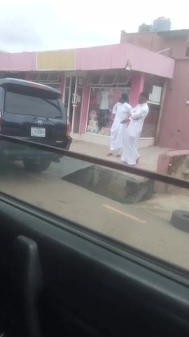 A black SUV is parked in front of a pink and brown building with storefronts. Two people dressed in white robes are standing on the sidewalk in front of the building, facing away from the camera. One person is bent over, appearing to be praying or examining something on the ground. The other person is standing upright. The SUV has a license plate that reads "SDF 316 GH". The storefronts appear to be selling clothing and other goods. The sky is overcast.