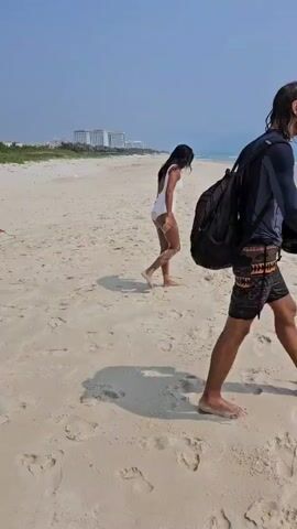 The video shows a wide-angle shot of a bright, sandy beach with a clear, pale blue sky above. The sea stretches into the distance, meeting the sky at the horizon with gentle waves lapping the shore. In the foreground, the sandy beach is covered with many footprints.

Initially, a woman with dark hair is walking away from the camera toward the water, wearing a white one-piece swimsuit. After a few steps, she pauses as another person, also in a white swimsuit and dark hair, turns to face her and takes a picture. Soon after, a third person, wearing dark clothing and a backpack, joins them running from the right side of the frame. All three then continue to move towards the water.