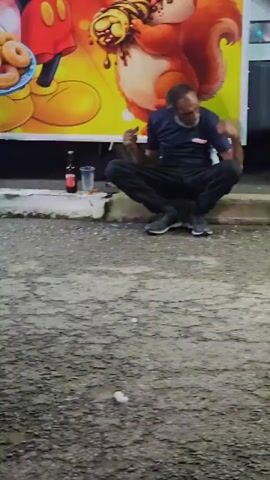 A man with a beard and dark clothing squats next to a wall with a colorful cartoon poster. He is holding a cigarette and looking at something in his hand. A bottle and a plastic cup are next to him on the ground. People walk past him in the foreground. The ground is paved and littered with debris.