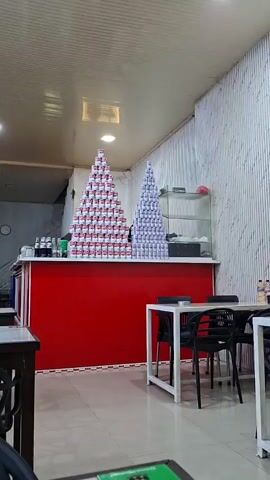 The video shows an interior shot of a casual dining establishment or cafe. In the center, a long red counter features two large, meticulously stacked pyramids made of beverage cans. The pyramid on the left consists of cans with red and white labels, while the one on the right is made of cans with blue and white labels. Behind the counter, a few beverage bottles and other items are visible, along with a glass display case to the right. In the foreground, several white tables with black chairs are arranged. The walls are light-colored, with a textured paneling on the right side. The ceiling is white with recessed lighting. The camera slowly pans slightly to the right, offering a wider view of the counter and its stacked cans.