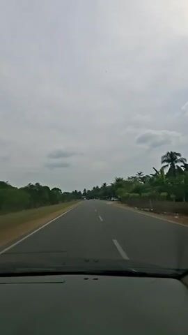 The video shows a POV shot from inside a car driving on a paved road. The road has a white dashed line down the center, indicating two lanes of traffic. On the left side of the road, there is a dirt shoulder and then a grassy field with scattered trees. On the right side of the road, there are more trees, some buildings, and parked vehicles, including a white car and several motorcycles. The sky is overcast with clouds. The video is short and does not show any notable interactions between people or objects.