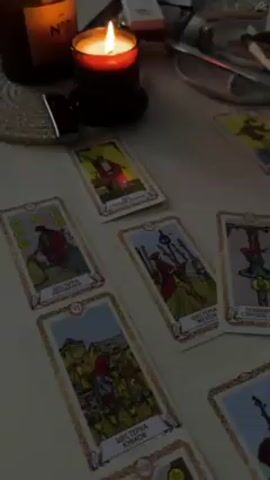 A high-angle shot shows a collection of Tarot cards spread out on a white surface around a lit candle. The Tarot cards are laid out with a thin gold border and a beige background. The cards display various images, including figures and symbols, with titles in what appears to be Russian.  A candle in a black holder sits to the left, emitting a warm glow. Beside the candle, various items including a lighter, a white box and a small ceramic dish with a smudge stick and other objects are visible in the background. The lighting is low, creating a dim and warm ambiance, emphasizing the ritualistic or divination context associated with Tarot reading. The surface appears to be a table or flat surface.