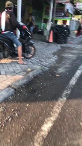 A person in a black jacket and blue shorts is sitting on a black motorcycle parked on a cobblestone sidewalk. The person is wearing flip-flops and a tan baseball cap. The motorcycle is facing left, with its front wheel turned slightly to the left. In the background, there is a building with a green awning and several stacked tires. The ground is wet and appears to be asphalt. The lighting suggests it is daytime, possibly late afternoon due to the long shadows.