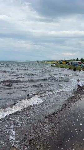 A short video clip shows a body of water with waves crashing onto a shore. The shore is made of pebbles and some grass. In the background, there are tents set up, suggesting a campsite. A person, possibly a child, is sitting near the water on the right side of the frame. The sky is cloudy.