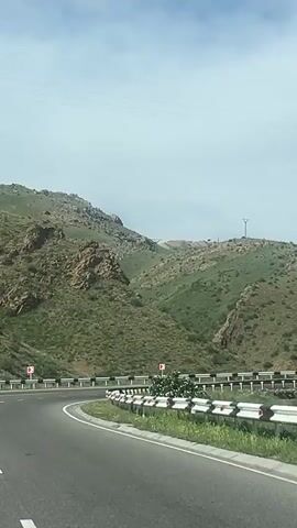 The video captures a daytime view from a moving vehicle driving along a winding asphalt road through a mountainous landscape, specifically in Yeraskh, Armenia. The road features clear white lane markings and is bordered by white and black guardrails. Two red and white chevron signs, visible on the right side of the road and further down on the left, indicate a right-hand curve ahead.

The surrounding environment consists of rugged mountains with a mix of rocky outcrops and green, grassy slopes covered in scrub vegetation. In the distance, on the mountain ridges, several power lines and tall power line towers (pylons) are visible against the sky. The sky is partly cloudy with patches of blue, suggesting fair weather. Some green bushes and foliage are present near the guardrails on the right side of the road. The overall scene depicts a scenic journey through a rural or semi-rural mountainous area.
