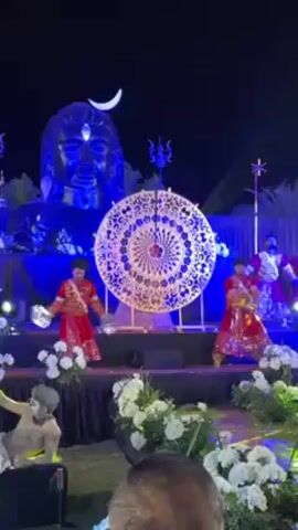 The video shows a nighttime performance on a decorated stage featuring a large sculpture of a deity with a crescent moon above its head. In front of the sculpture, a large, circular, intricate metal design serves as a focal point. Two dancers in red and gold costumes with small hand cymbals are performing on the stage.  Additional performers are visible, some holding poles, and there is another individual with painted skin standing to the side. The stage is decorated with white flowers and blue lighting. The audience can be seen in the foreground. The setting appears to be an outdoor event.