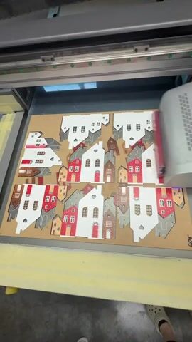 The video shows a large format printer operating on a flatbed, printing a series of small house cutouts. The cutouts are made of a tan material, possibly cardboard or wood, and are arranged in a grid-like pattern. The houses are designed with different colored roofs (red and grey) and windows. Some of the houses have heart-shaped windows. The printer head is moving back and forth across the material, and the cutouts are visible beneath it. The environment appears to be a workshop or manufacturing facility, with industrial equipment and lighting. The video is shot from a top-down perspective.