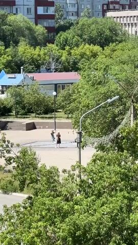 The video pans across a scene with buildings and trees in the background. In the foreground, a fountain is active with water spraying upwards. Two people are visible near the fountain. One person is standing and appears to be holding something, possibly a phone. Another person, dressed in dark clothing, is walking away from the fountain. The sun is shining, casting shadows, indicating it is daytime.