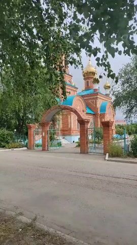 A video shows a brick church with golden domes and blue roofs. A brick archway with metal gates stands in front of the church. Trees with green leaves are visible in the foreground. The camera pans and zooms in on the church and its domes.