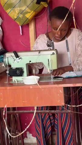 A young woman is seated at a sewing machine, working on a piece of fabric. The sewing machine is a light green vintage model, placed on a wooden table with a red and blue striped base. A white bowl sits next to the machine. The woman is wearing a light pink shirt with lace sleeves, and a measuring tape hangs around her neck. She is focused on her work, her hands guiding the fabric under the needle. The background includes a pink wall and a bag hanging from a wooden beam. There are also string lights around the room, adding a warm glow to the scene.