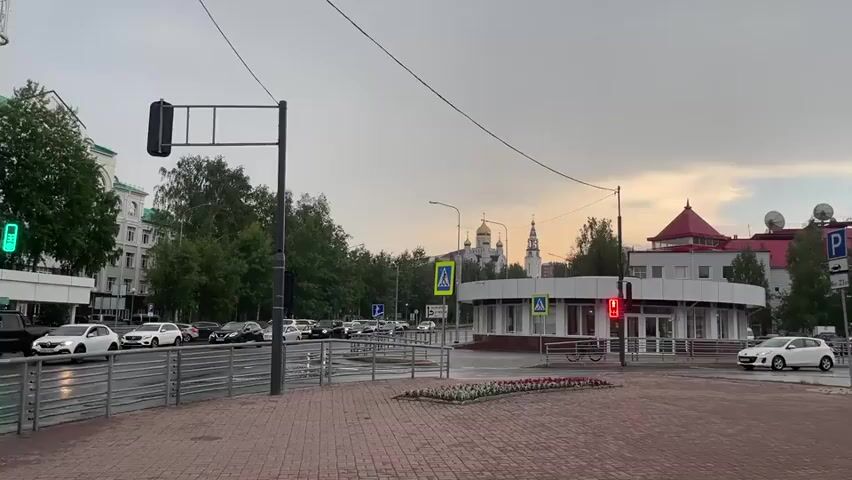 This media file depicts an outdoor urban scene, likely a street intersection in Khanty-Mansiysk, Russia, captured during the late afternoon or early evening. The sky is overcast with a hint of warm light peeking through, suggesting a possible sunset or clearing storm. The weather appears to be damp, as indicated by the wet road surface and the reflections on it.

The primary subjects are a street and its surroundings. Cars are visible, driving on the road and parked along the side, indicating normal city traffic. No people are clearly discernible in the foreground or mid-ground, though there might be individuals on the sidewalks of the distant buildings. The architecture includes multi-story buildings, some with classic European styles, and a distinct circular building with a white facade and a red-roofed extension. In the background, a church with golden domes stands prominently against the sky, a common landmark in many Russian cities.

Several objects are notable: traffic lights, a pedestrian crossing signal displaying green, street signs, a traffic pole with a black signal box, and a metal railing separating the sidewalk from the road. A flowerbed with colorful blooms is situated in the center of the intersection.

The activity is the flow of traffic and the general ambiance of a city transitioning from daytime to evening. There are no overt signs of any specific event taking place, rather it’s a depiction of everyday urban life.

Notable details include the Russian text on the street signs: "ул. Гагарина" (Gagarin Street) with an arrow pointing upwards, and "ул. Мира" (Peace Street) with an arrow pointing to the right. This confirms the location as Khanty-Mansiysk. The overall mood is calm and slightly subdued, possibly due to the overcast weather.