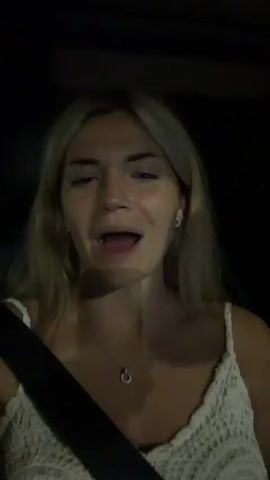 A young woman with blonde hair is in the passenger seat of a car at night. She is wearing a white top and a seatbelt. She is looking at the camera and talking. The lighting is dim.