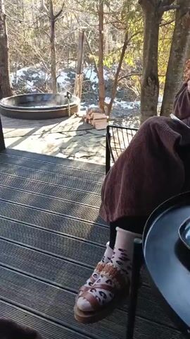 The video depicts a person relaxing on a wooden deck, showcasing a hot tub steaming in the background amidst a forest landscape. The view includes glimpses of remaining snow patches. The person initially has on socks and clogs, then switches to flip-flops as they stretch their legs out. The deck is made of grooved planks, and beyond it lie trees and a partial view of the forest floor. The person seems to be enjoying a tranquil moment outdoors.