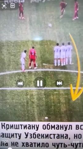 This is a top-down aerial shot of a soccer match. A player in a red jersey is attempting to score a goal. He dribbles the ball past two defenders in white jerseys. As he approaches the goal line, several players in white jerseys attempt to block him. The camera is focused on the action, and the ball is visible near the goal line. The grass field has white markings. There are also control buttons for a video player overlaid on the bottom of the screen. The text at the bottom of the screen is in Russian.