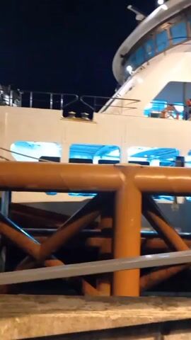 At night, a medium shot shows a white ferry docked at a pier.  A white metal gangway with "SEAFR" painted on the side connects the ferry to the pier. A few people are standing on the deck of the ferry near the entrance to the gangway.  A man in a white hardhat and an orange safety vest walks on the gangway. The pier has an orange pipe support structure and metal guardrails. The night sky is dark.