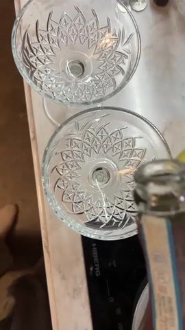 Here's a concise description of the video:

The video shows a person pouring Prosecco into two crystal coupe glasses on a marble countertop. The glasses feature intricate geometric designs at the bottom. The bottle of Prosecco has a burgundy label with blue and gold accents.  A partially peeled banana is visible near the glasses. The countertop is situated above a black stove top. A small part of the person's foot is visible on the lower left of the frame.
