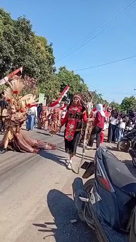 The video shows a parade on a paved street on a sunny day. In the foreground, a person in traditional costume, including a headdress, red shirt, black pants, and white sneakers, walks while holding a staff. The costume has intricate beadwork and embroidery. To the left, another person in an elaborate, ornate costume with feathers and a long, flowing skirt is visible. Behind them, other participants in traditional attire and carrying flags, including the Indonesian flag, walk along the street. Spectators are lining the street, some on motorcycles parked on the side. Trees and buildings are visible in the background. The sky is clear and blue.
