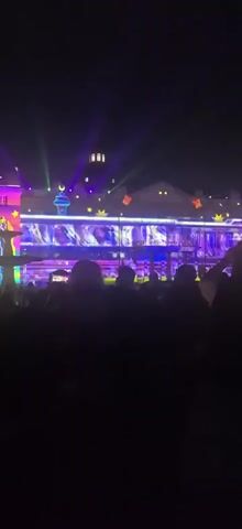 A dark night scene with a crowd of silhouetted people facing a building illuminated by vibrant light projections. The building's facade is covered in colorful, abstract patterns, some resembling aurora borealis, and celestial motifs like crescent moons and stars. Green, purple, and pink lights beam upwards from behind the crowd and from the building itself. The projection on the building's lower level appears to be a continuous sequence of shifting, colorful images. Several people in the foreground are holding up their phones, likely recording the display.