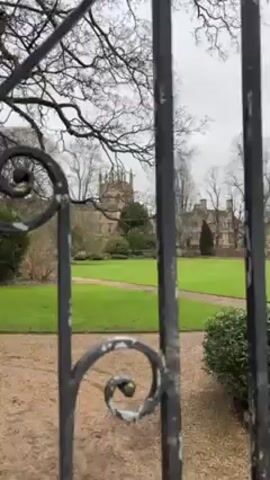 The camera looks through an ornate wrought iron gate, with scrollwork designs, at a building in the distance surrounded by a green lawn and trees. The gate is painted black and shows signs of age. The building in the distance has a distinctive spire. The sky is overcast. A gravel path is visible inside the gate. The trees are bare, suggesting it is winter or early spring. There are shrubs and other greenery inside the gate. The camera moves slightly.
