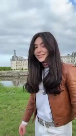 The video takes place outdoors, in front of what appears to be the Château de Chambord in France. The sky is overcast. The subject is a young woman with long dark hair. She is wearing white pants, a white t-shirt, a brown jacket, and a patterned scarf around her neck. She is standing on a grassy bank next to a body of water that reflects the castle. She first smiles at the camera, then "dabs," then performs a cartwheel. After the cartwheel, she whips her hair back and then fixes it with her hands.