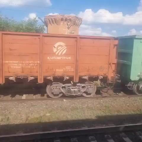 The video shows a side view of multiple train cars moving along a track. The train cars are various colors, including green and light blue, and some have Cyrillic writing and numbers visible on them. The background features a blue sky with scattered white clouds and some trees and vegetation. The train appears to be in motion, indicated by the slight blur and the changing perspective of the scenery. The camera is positioned at a level that captures the side of the train cars and the track below. The lighting suggests it is daytime, likely with sunny conditions.