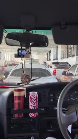 The video shows the interior of a car looking forward, through the windshield. The car is on a ferry, as indicated by the rows of other vehicles visible in the background. The dashboard of the car is in the foreground, with a steering wheel on the right. A dash cam is attached to the rearview mirror, and prayer beads hang from it. Two drinks, a can of Coca-Cola and a red can labeled "LOHAS", are in a holder on the dashboard. The video is static, with no apparent movement.