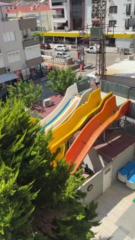A drone or elevated camera provides a bird's-eye view of a resort or hotel complex. In the foreground, a large, lush green tree partially obstructs the view. Beyond the tree, a swimming pool filled with bright blue water is visible, with several people swimming and a smaller pool adjacent to it. Sun loungers with orange cushions are arranged around the main pool. In the background, a street with cars and buildings can be seen, including a gas station and apartment buildings. Colorful water slides are also visible, indicating a recreational area within the complex. The sky is clear and blue, suggesting a sunny day. The camera pans across the scene, showing different sections of the resort and its surroundings.