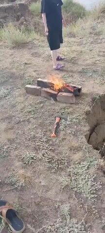 A person stands on a dirt cliff overlooking a body of water. In front of the person is a campfire built with bricks. An axe lies on the ground near the campfire. The person walks away from the campfire and towards the edge of the cliff.