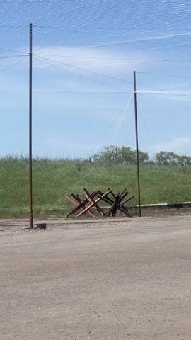 The video shows a daytime outdoor scene with a dirt road in the foreground, two tall metal posts and a "Czech hedgehog" anti-tank obstacle in the middle ground, and a grassy field with some trees in the background.  Above the scene is a grid-like net or screen that obscures the blue sky. There is no visible human presence or activity in the video.