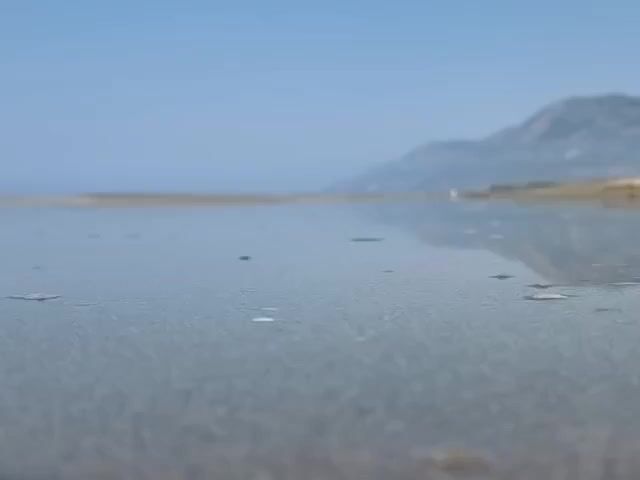 A low-angle shot shows a very shallow body of water covering sand. The water is calm with a few small bubbles visible on the surface. In the background, a blurred landscape of sand and mountains under a clear blue sky is visible. The scene appears to be a beach or a tidal flat on a sunny day.