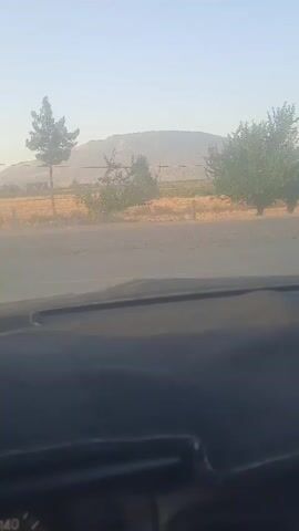 A first-person perspective video captures the view from inside a vehicle. The camera pans slightly to the right, revealing the dashboard and a portion of the steering wheel. Outside the windshield, a hazy landscape unfolds with a prominent hill in the distance, flanked by trees and dry, yellowish fields. The sun is visible in the sky, casting a bright glare. The video then shifts focus to the side mirror, which reflects a truck traveling on the road behind. The interior of the car appears to be dimly lit, while the exterior is bathed in sunlight. The overall impression is of a car traveling through a rural or semi-rural area during daylight.