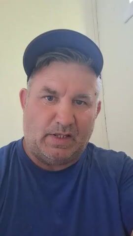 A man wearing a blue t-shirt and a dark blue baseball cap is looking directly at the camera. He has short graying hair, stubble on his face, and light-colored eyes. He appears to be indoors with a light-colored wall in the background.