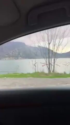 The video shows a car driving along a road with a scenic view. Initially, the view from the passenger side window reveals a body of water with mountains in the background. The water has some floating objects arranged in rows. As the video progresses, the perspective shifts to the driver's view. The road ahead is lined with sparse vegetation, and towering mountains dominate the landscape. The sky above is overcast, adding to the dramatic atmosphere of the scene. The road is paved with white lane markings.