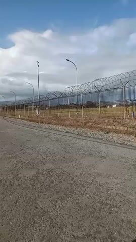 The video shows a person riding a motorcycle on a dirt road next to a tall fence topped with barbed wire. The fence is long and appears to enclose a large area. There are streetlights along the fence. The sky is blue with some clouds. The land beyond the fence is flat and appears to be undeveloped. The video is shaky, indicating it was filmed while moving.