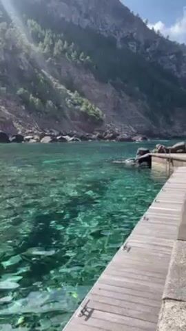 The video shows a serene view of clear turquoise water from a wooden dock or pier. The water is so transparent that the bottom of the bay is visible, revealing rocks and the subtle motion of the water. In the background, a steep, rocky hillside covered with trees rises sharply. The sun's rays filter through the scene, creating a play of light and shadow on the water's surface. The camera seems to be positioned slightly above the water, giving a clear view of the surroundings. There appear to be a few people on the rocks in the middle distance. The overall impression is one of natural beauty and tranquility.