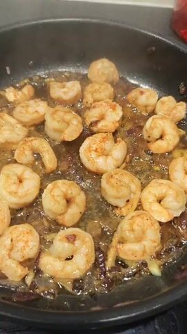 A static shot shows a pan on a stovetop, containing shrimp sauteing in oil with small pieces of red onion. The shrimp are pink, opaque, and curled. Steam rises from the pan.
