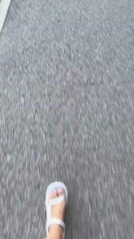 The video shows a first-person perspective of someone walking on an asphalt surface. The person is wearing white sandals. A white line is visible on the left side of the asphalt. Further down the path, a rectangular metal grate is visible embedded in the asphalt. The overall impression is of a person walking along a sidewalk or street.