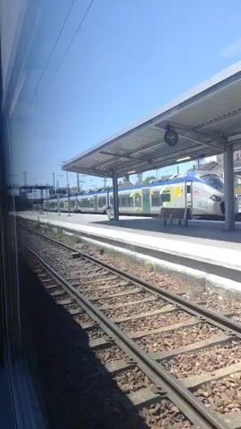 The video shows a train station with a train stopped at the platform. The camera is inside another train, looking out the window. The train at the station is a modern electric multiple unit with a silver and blue livery. It appears to be a regional train. The station platform is covered by a canopy, and there are benches and a clock visible. The tracks in the foreground are ballast track. The sky is clear and blue. The video is taken from a moving train, as evidenced by the slight blur and the changing perspective of the scenery.