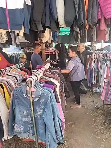 The video shows an outdoor market stall selling used clothing. Several racks of clothing are visible, hung from metal poles and a makeshift canopy. The clothing includes a variety of jackets, shirts, and pants in different colors and styles. Three people are interacting at the stall. One person, a man wearing a blue shirt and a cap, appears to be attending the stall. Two women are browsing the clothing. One woman, wearing a hijab and a dark jacket, is looking at some items. The other woman, wearing a light-colored jacket and dark pants, is standing next to her. The ground is dirt, and there is some dry vegetation visible. The overall impression is of a casual, open-air market.