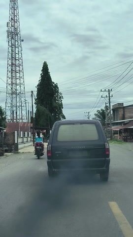 The media file depicts a street scene in Kutapaya, Indonesia, under an overcast sky. The camera is positioned inside a vehicle, looking out at the road.

The primary subjects are two people riding motorcycles and a dark-colored van. The first motorcycle, closer to the camera, carries two individuals. The rider wears a light blue shirt and a dark helmet, while the passenger has long dark hair and wears a light-colored garment. The second motorcycle, further ahead and to the left, also appears to have two people on it, one with long dark hair. The van is in front of the motorcycles, traveling in the same direction. It is a dark color with tinted rear windows. Faint text is visible on the rear window of the van.

The scene is set on a paved road with a yellow lane marker visible. To the left of the road, a large, mature tree with dense foliage dominates the landscape, its branches extending towards the sky. Behind the tree, a tall, lattice-like telecommunications tower stands prominently. Further down the road, power lines are strung between utility poles, typical of a semi-urban or rural area. The background reveals buildings with roofs and some vegetation, including palm trees, suggesting a tropical climate.

The activity shown is travel along a road, with the motorcycles seemingly following the van. The overcast sky suggests it might be daytime, possibly late morning or afternoon, but the lack of strong shadows makes it difficult to pinpoint precisely. The weather appears to be cloudy, perhaps humid. The overall mood is neutral, capturing a mundane moment of transit. The visible text on the van's rear window is too indistinct to be read.