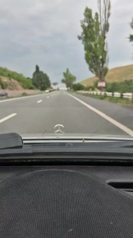 The video shows a view from inside a car driving on a highway. The dashboard is visible in the foreground with the Mercedes-Benz logo on the hood. The road ahead is a two-lane highway with a white dividing line. On the left side of the road, there are green hills and trees. On the right side, there are also trees and a guardrail. The sky is cloudy. Several cars are visible on the road, including a black sedan in front of the camera car and a white car on the left lane. The black sedan is in the right lane and appears to be slowing down. The video captures the moment the black sedan passes by.