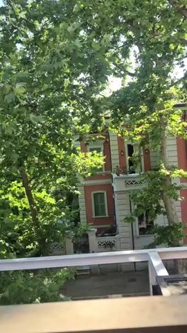 The video pans across a balcony railing, then moves to show a tree-lined street with a red and white building. The building has several windows and a balcony. The camera then pans up to show the sky and more trees.