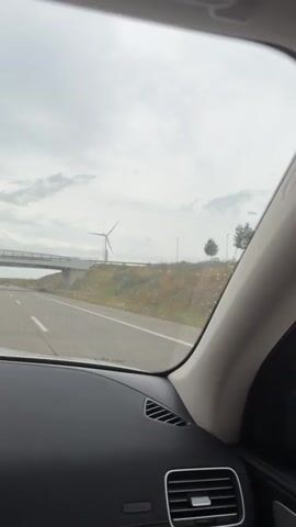 The video is taken from inside a car, looking out the front windshield and side window. The car appears to be on a highway, with a white lane marker visible on the road. In the distance, there is a bridge and several wind turbines. The sky is overcast and there are some raindrops visible on the windshield. The side mirror of the car is also visible, showing a reflection of the car and the passing scenery. The overall impression is a view of a highway during a cloudy day.