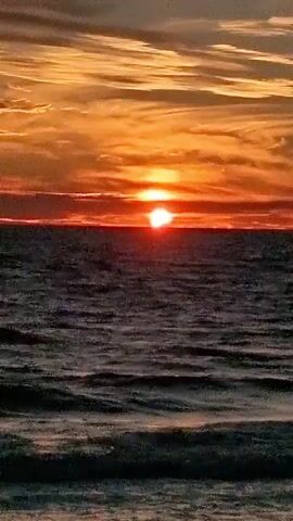 This is a static, vertical video of a sunset over the ocean. The sun is a bright orange orb on the horizon, partially obscured by clouds. The sky is a vibrant mix of orange, yellow, and red. The ocean is dark and choppy, with small waves. The bottom of the frame shows a dark shoreline, possibly sand. There are no people or objects visible in the video.