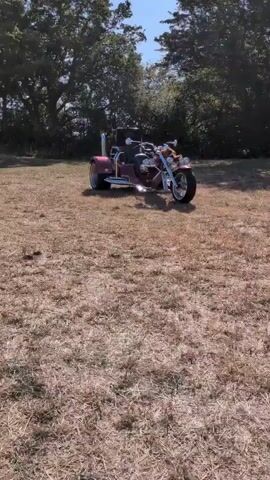 A trike is parked on a dry grassy field, with trees in the background under a clear blue sky. The trike is red and has chrome accents. The camera pans slightly to the left.