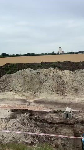 The video shows a construction site with a large pile of dirt in the foreground. A body of muddy water is visible in a depression in the dirt. In the background, there is a field of dry, golden crops and a tall, white building. The sky is overcast. The video appears to be taken from a moving vehicle, possibly a car, as indicated by the slight blur of the image. There are no people visible in the video.